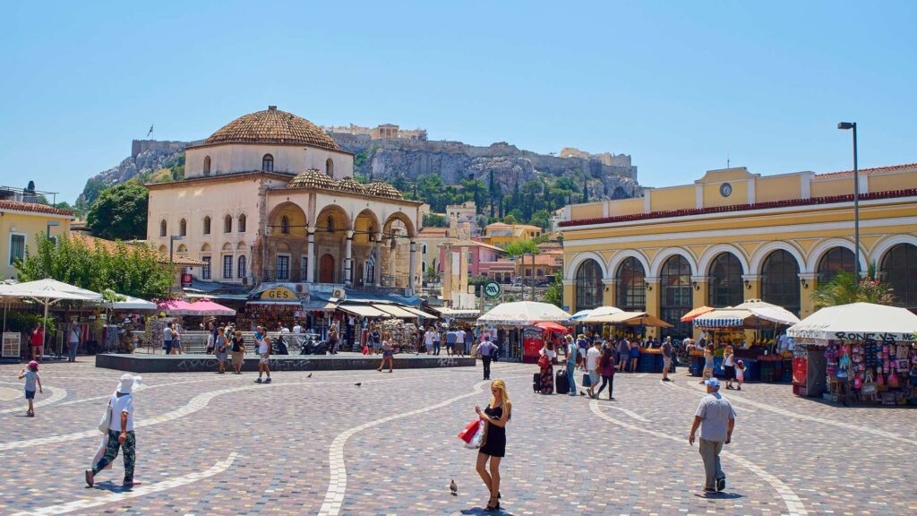 The square bustles with shops and people under a clear sky. On the left stands an old domed building, while a large yellow one is on the right. An ancient hilltop structure graces the background, leaving visitors to ponder how many days in Athens they might need.
