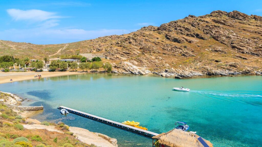 A serene beach in Naoussa, Paros, with clear turquoise water, a rocky hill in the background, a dock, and a few boats on the water under a clear blue sky.