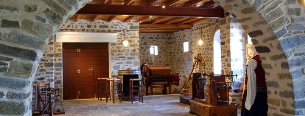 The interior of this traditional stone room in Naoussa, Paros features wooden ceiling beams, wooden doors, and stools. Historical artifacts are on display, including a mannequin dressed in period clothing, beautifully capturing the essence of this enchanting Greek locale.
