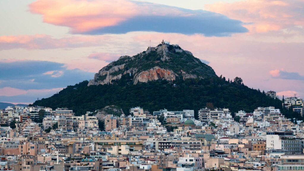 Marvel at the breathtaking view of Lycabettus Hill surrounded by urban buildings at sunset, with a partly cloudy sky. Witness this beauty as you ponder how many days in Athens you'll need to fully embrace its charm and history.
