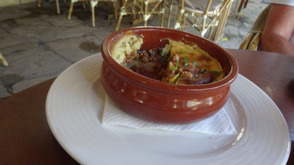 A partially eaten dish in a brown ceramic bowl on a white plate, resting on a napkin, offers a glimpse of Greek street food flavors. The dish features mashed potatoes and minced meat, delicately garnished with herbs.