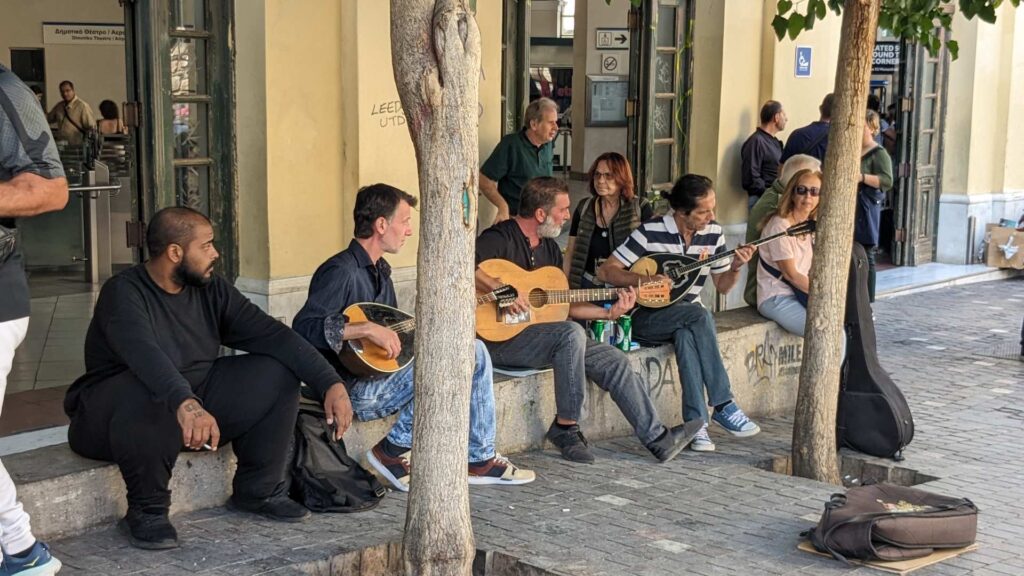 Street musicians play guitars and bouzoukis as people gather on a city sidewalk, soaking in the vibrant atmosphere of Athens. Trees and a building create a picturesque backdrop, making every moment feel like part of an adventure that begs the question: how many days in Athens are truly enough?.
