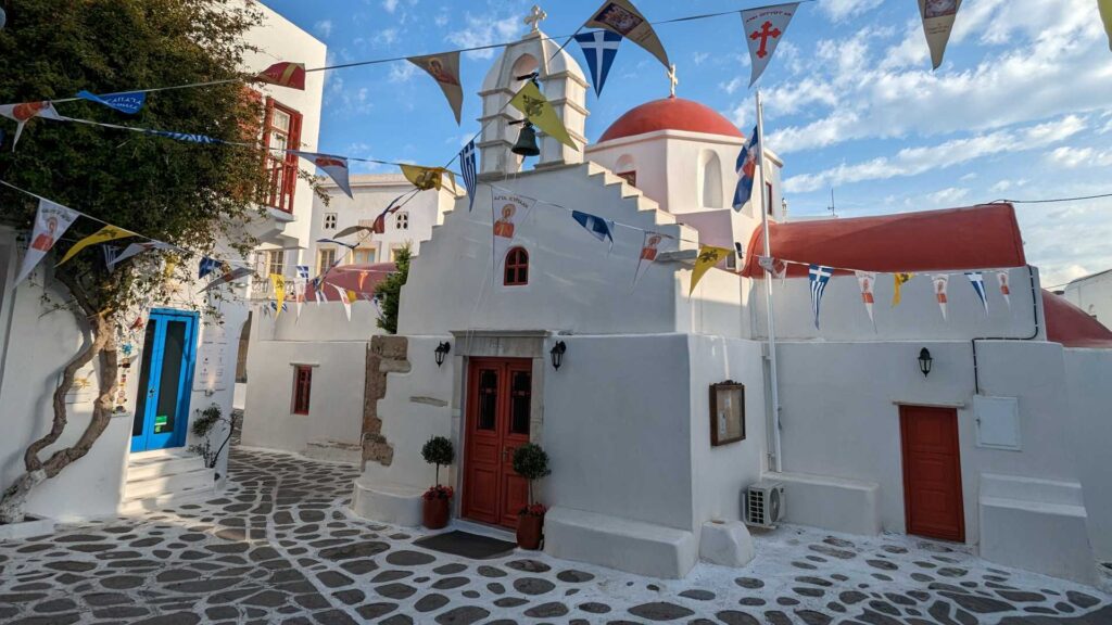 A small white church with a red dome, decorated with flags, graces the charming Naxos landscape. It's nestled in a cobblestone courtyard and surrounded by white buildings under a brilliant blue sky.