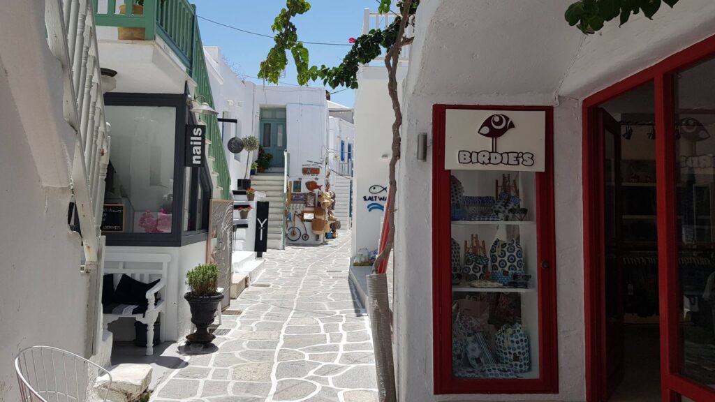 Narrow cobblestone street in Naoussa, Paros, lined with white buildings, small shops with signs, and vibrant green plants under a clear blue sky.