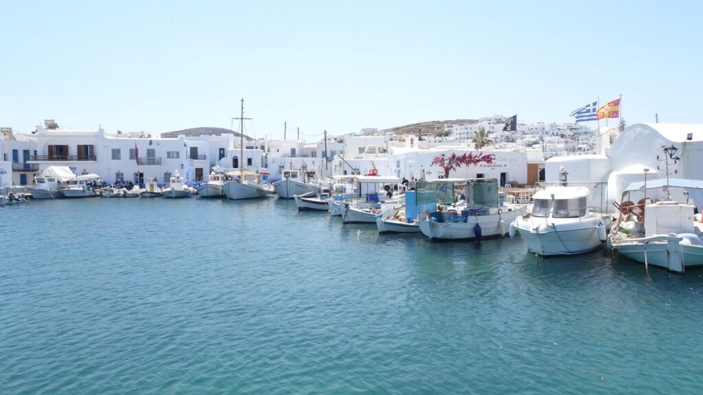 Boats are docked along the scenic harbor of Naoussa, Paros, with white buildings in the background under a clear blue sky.