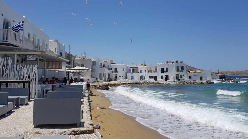 A seaside cafe in Naoussa, Paros, where people dine on a sandy beach beside white buildings under a clear blue sky, with seagulls gracefully soaring overhead.