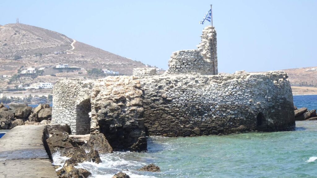 An ancient stone castle ruin by the sea features a Greek flag atop, with Naoussa's picturesque hills and scattered white buildings of Paros as its stunning backdrop.