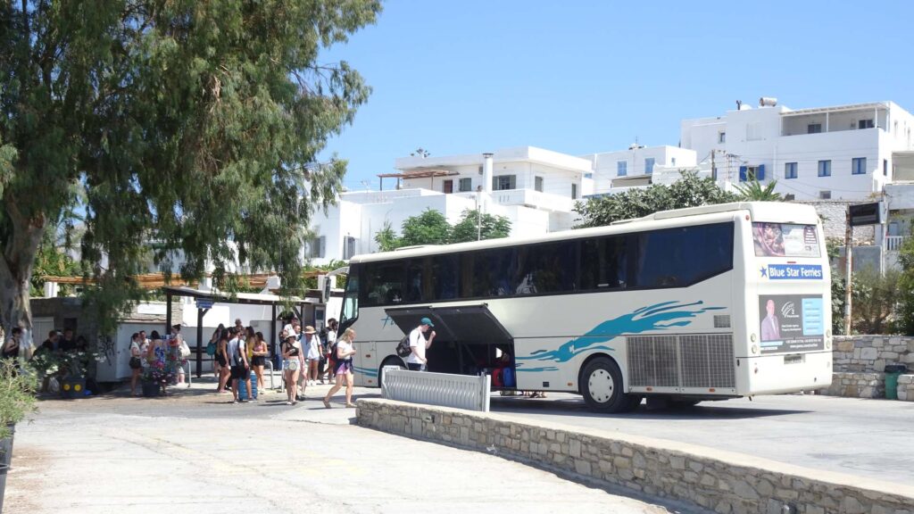 A tour bus parked on a sunny street in Milos, with passengers boarding and disembarking, surrounded by white buildings and trees.