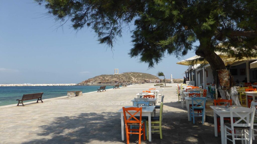 The seaside promenade in Naoussa, Paros features colorful chairs and tables next to the water. A tree provides shade, while a small hill with a quaint structure is visible in the background, adding to the picturesque charm.