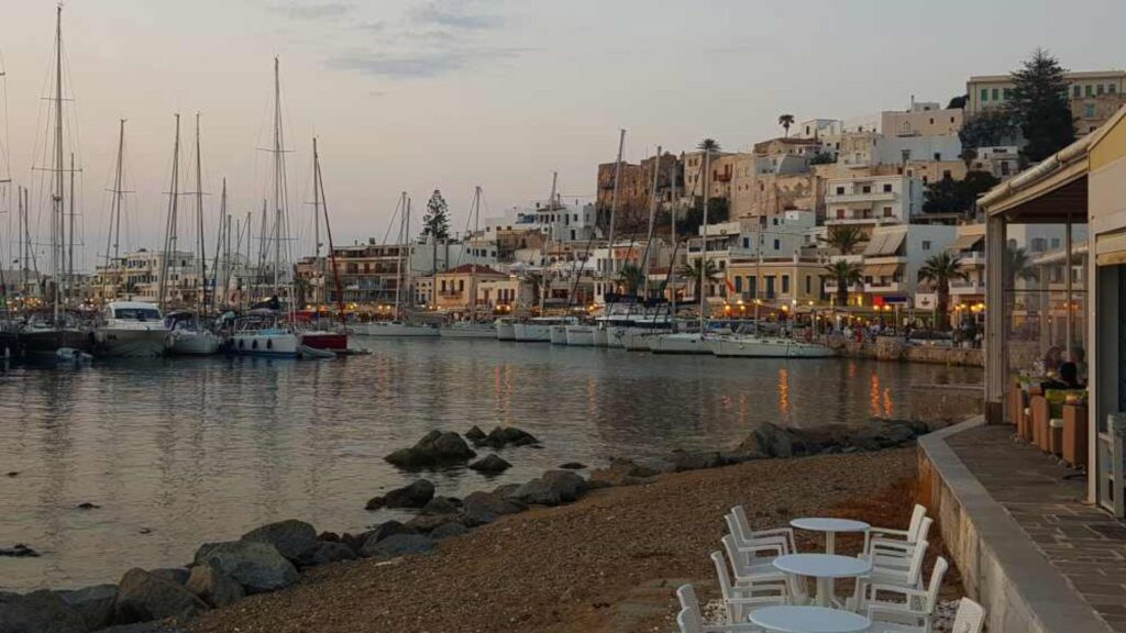At sunset, a serene harbor in Naxos reveals sailboats docked peacefully, with charming buildings perched on the hillside. In the foreground, an empty patio with tables and chairs offers a quiet spot to take in the tranquil beauty of the scene.