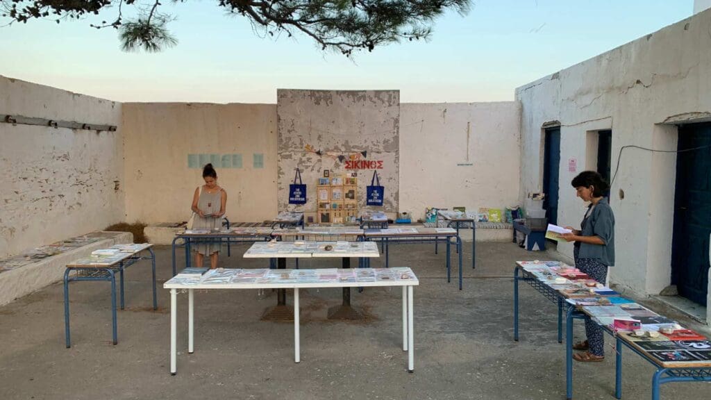 Two people browse books and magazines on tables in an outdoor space with white walls and a tree overhead, capturing the serene essence of Sikinos Island.