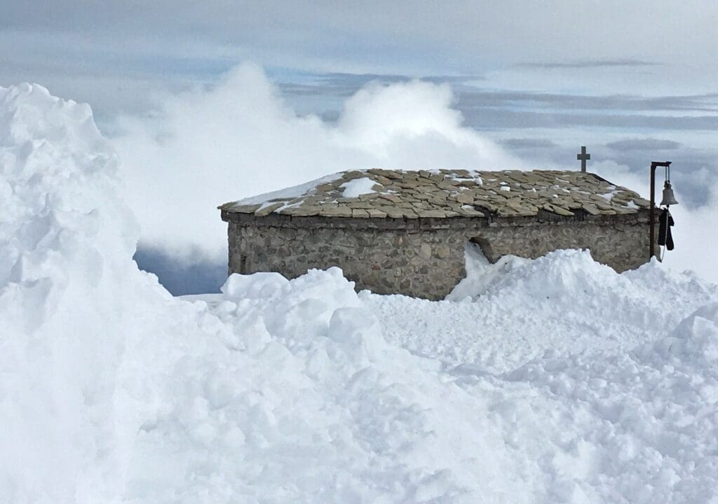 A stone chapel nestled in a snow-blanketed landscape offers a serene backdrop for those exploring the best Greek ski resorts on a cloudy day.