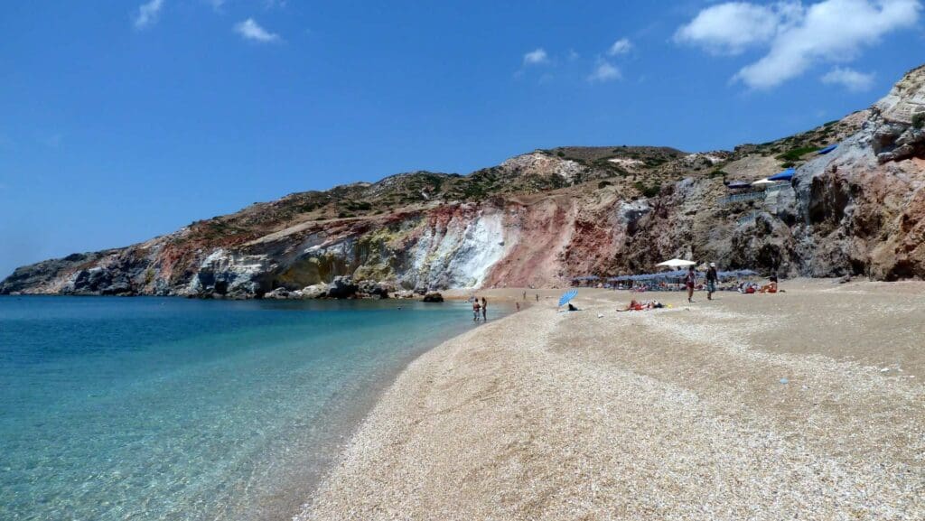 A sandy beach on Milos with scattered people under umbrellas, clear turquoise water, and rocky cliffs in the background under a blue sky makes it one of the must-see spots.