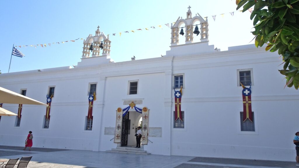 Nestled in the heart of Paros, this white building with three bell towers stands proudly, adorned with flags and banners. People gather eagerly at the entrance, perhaps drawn by tales from their Island Guide, eager to explore its storied past.