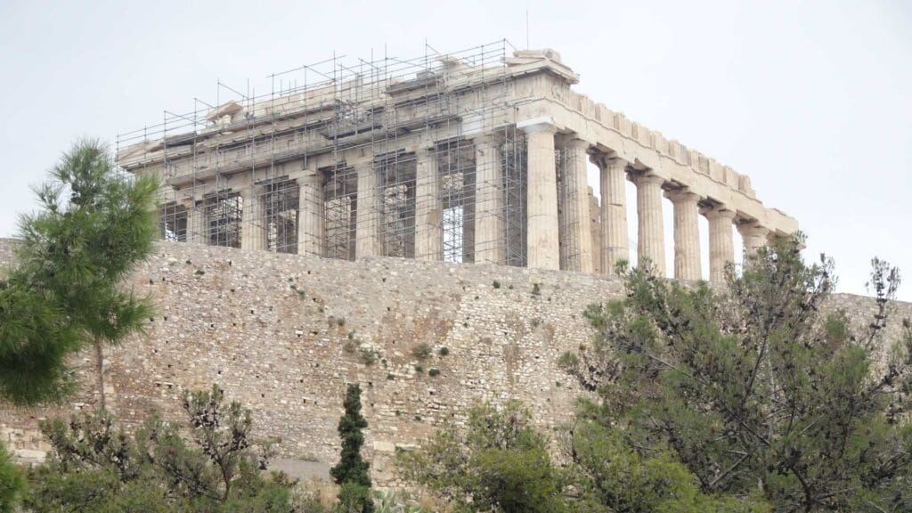 The Parthenon in Athens, Greece, partially covered in scaffolding, stands atop a stone wall surrounded by trees, offering a stunning highlight for any acropolis self-guided tour.