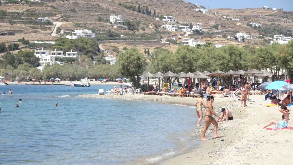 A beach scene on Naxos with people relaxing under straw umbrellas, swimming, and sunbathing. Hills with charming houses dot the background.