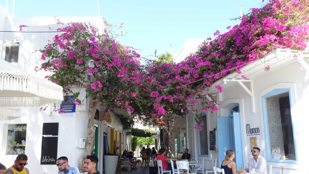 Amidst the charming white-walled town, travelers enjoy leisurely moments at outdoor cafes beneath bougainvillea-clad arches. The bright blue sky completes this idyllic Greek island-hopping scene.