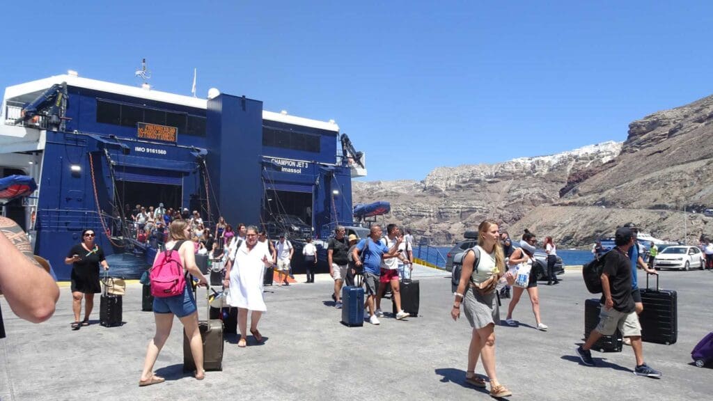 A crowd disembarks a large blue ferry at the Santorini ferry port, stepping onto the dock with rocky cliffs as their backdrop. Under the clear blue sky, passengers carry luggage, eager to explore.