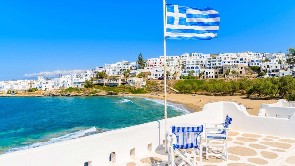 The Greek flag waves on a terrace in Naoussa, Paros, overlooking a sandy beach and turquoise sea with white buildings scattered on the hillside under a clear blue sky.