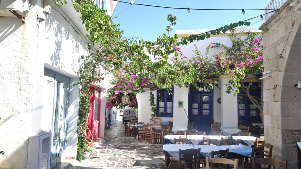 Quaint outdoor cafe with wooden tables and chairs on a narrow, cobblestone street, surrounded by white buildings and vibrant bougainvillea vines—a must-see spot to relax after you discover Milos.