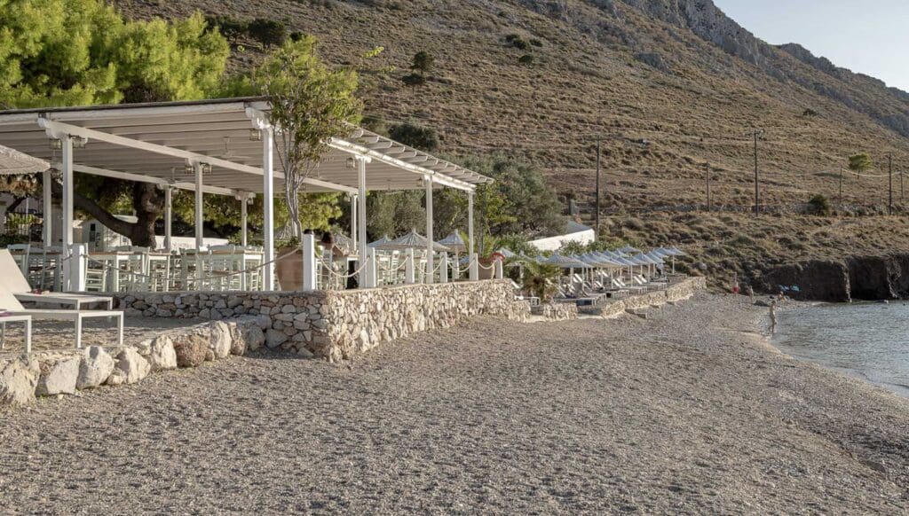 A stone-walled beachside seating area with white furniture under a pergola overlooks the pebbly shoreline and calm sea of Hydra beaches, with rocky hills in the background.