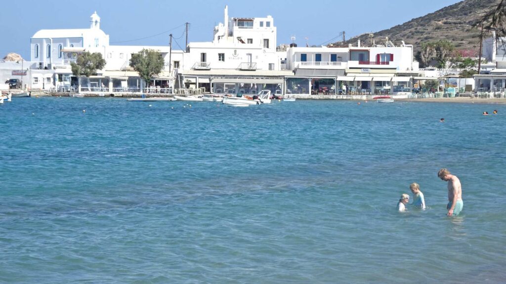 A couple stands with a child in the shallow, clear blue water of Paros. A row of white buildings graces the shoreline in the background, resembling a scene from an idyllic island guide.