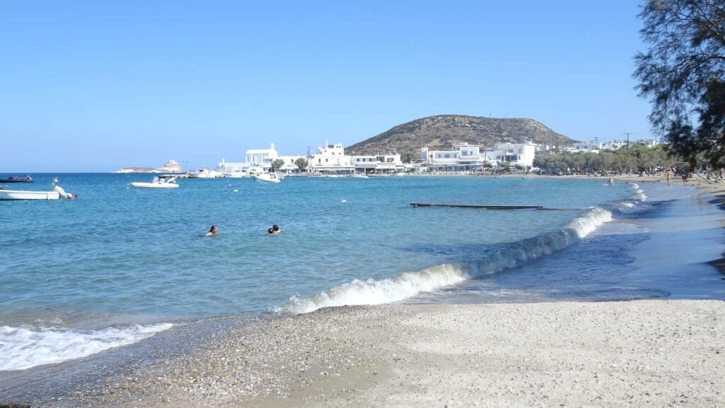 Discover a tranquil beach with clear blue water, where a few swimmers enjoy the serene surroundings and boats drift lazily in the distance. Nestled among Milos Beaches, this must-see spot features buildings and a hill under a crystal-clear sky.