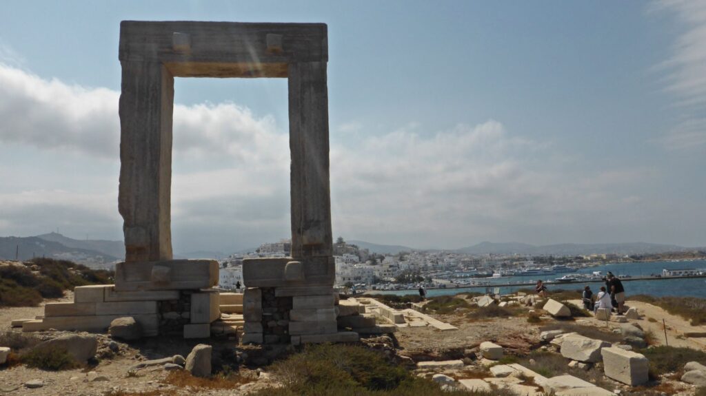 An ancient stone gateway on a hill overlooking the coastal city and harbor of Naxos, with scattered ruins nestled under a partly cloudy sky.