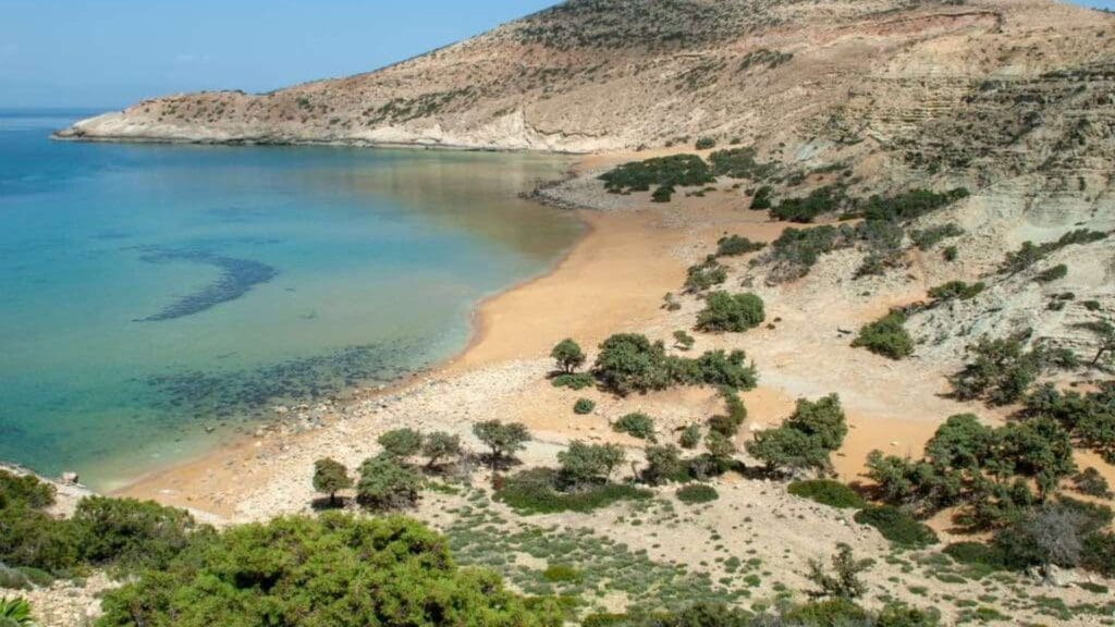A secluded beach with golden sand and clear turquoise water, reminiscent of the picturesque Heraklion beaches, is surrounded by rocky hills and sparse greenery.