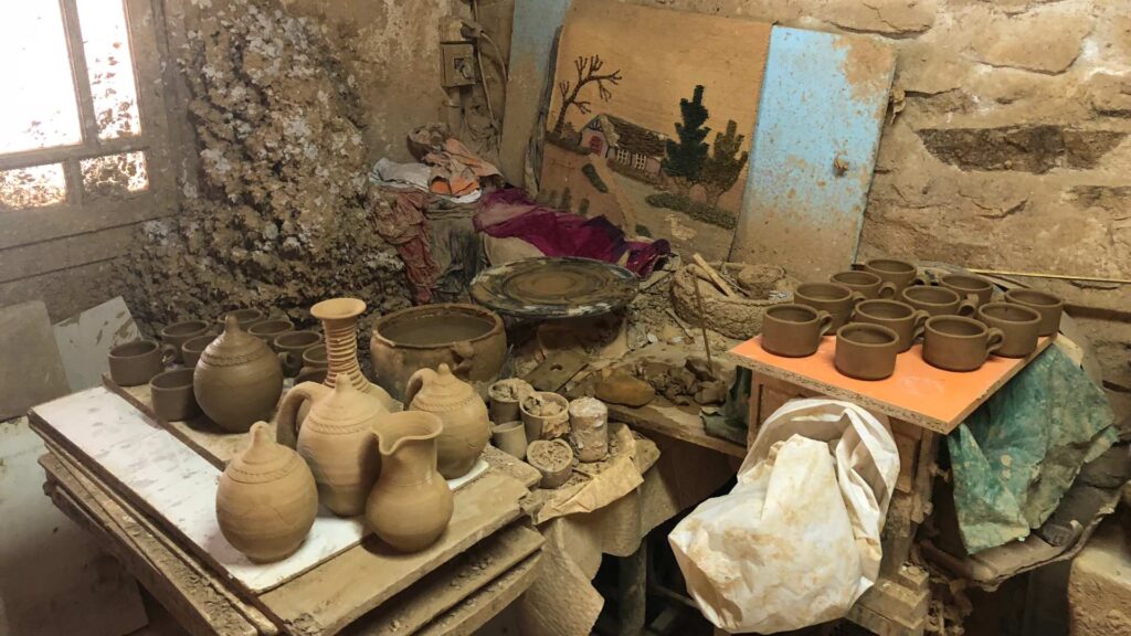 A cluttered pottery workshop on Paros, with various clay pots, jugs, and bowls drying on tables. A tapestry hangs on the wall among old tools and materials, capturing the island's artistic spirit.