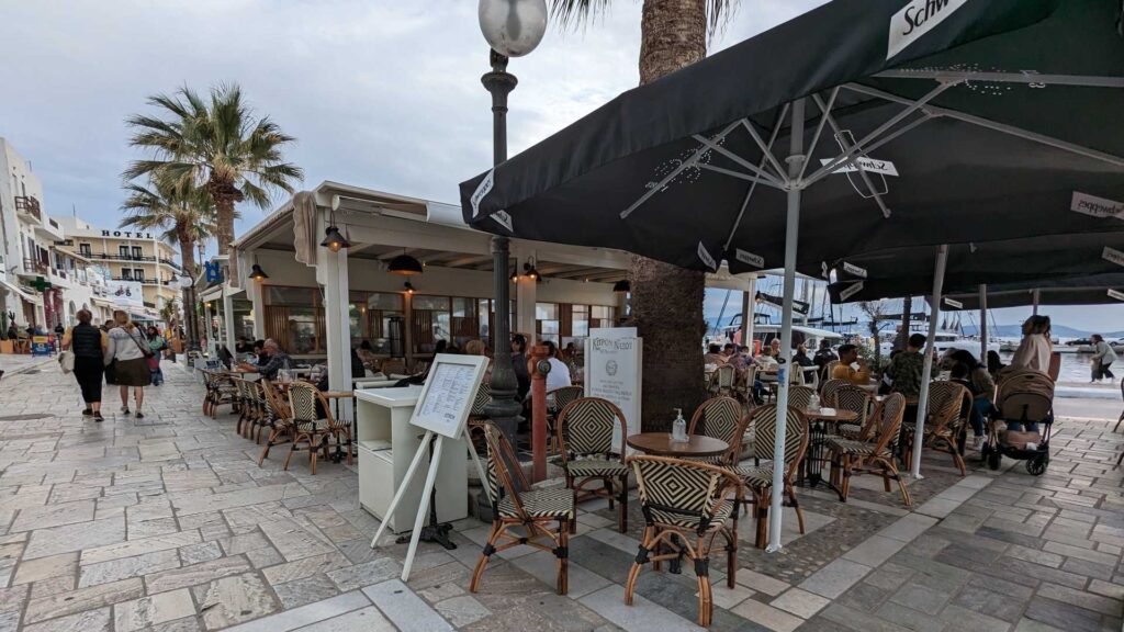 The outdoor café by the waterfront, reminiscent of Naxos charm, features people seated under large umbrellas with palm trees swaying gently nearby. A menu stand welcomes visitors at the entrance while the overcast sky adds to its cozy allure.