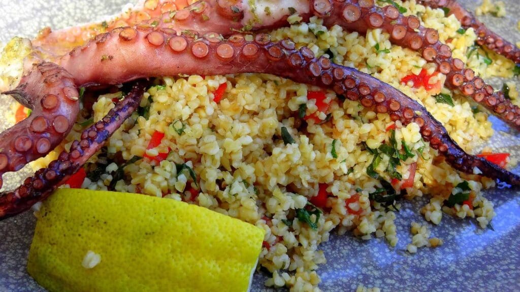 Grilled octopus tentacles, a Greek street food delight, served on a bed of bulgur with herbs, red peppers, and a lemon wedge on the side.