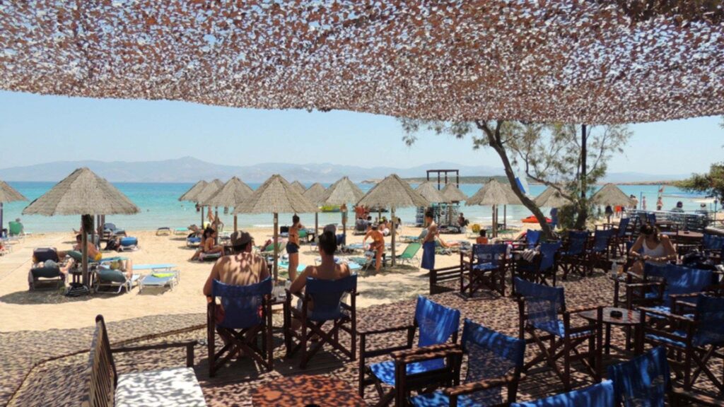 People relax under a shade canopy by a beach in Naoussa, Paros, adorned with sun loungers, umbrellas, and trees. The stunning sea and mountains create a breathtaking backdrop.