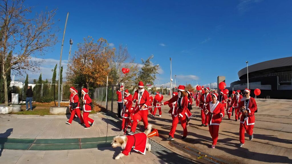A festive parade in Athens features a group of people dressed as Santa Claus strolling down the sidewalk with red balloons, while a dog in a Santa outfit prances by on a leash. This lively scene captures the essence of Christmas spirit among the top things to do in Athens during the holidays.