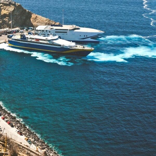 Ferries docked at the bustling Santorini ferry port, with several vehicles lined up along the pier and waves weaving patterns in the water. Rocky cliffs surround the area, adding to its scenic charm.