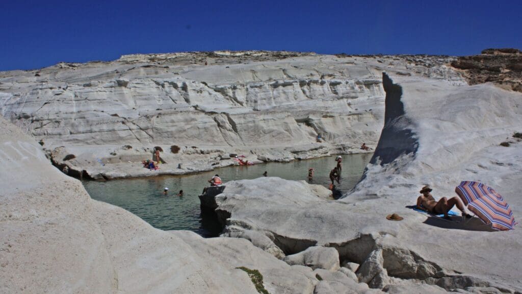 People relax and sunbathe on the moonlike paradise of white rock formations at Sarakiniko Beach in Milos, beside a natural turquoise pool under a clear blue sky.