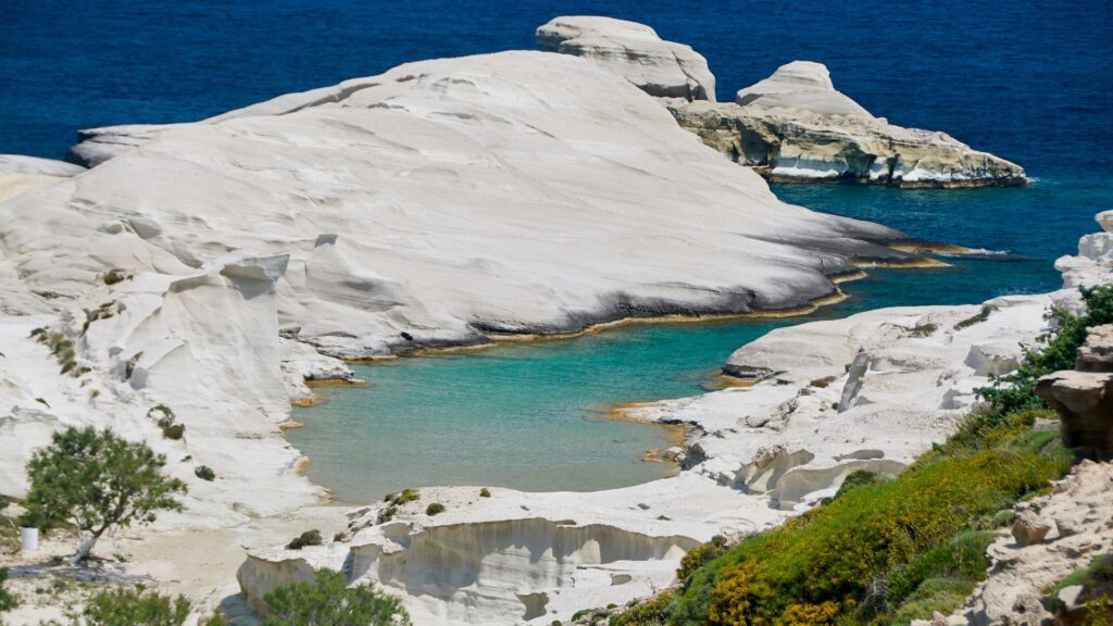 White rocky cliff formations on the island of Milos surround a turquoise inlet, with the deep blue sea in the background and lush greenery in the foreground.