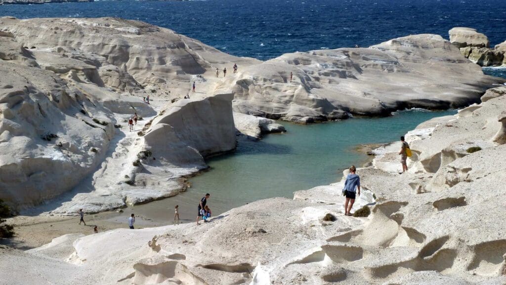 Under a blue sky, visitors explore the rocky white cliffs of Milos and a small, clear turquoise inlet, which are among the must-see spots on this enchanting island.