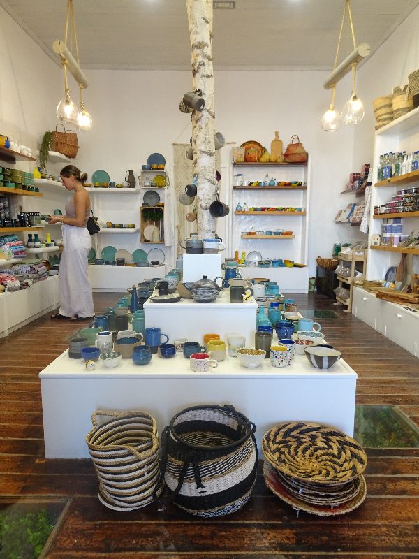 A person browses in a store featuring pottery, baskets, and various home goods reminiscent of the charming styles found on Paros. These treasures are thoughtfully displayed on shelves and a central table, capturing the essence of island life.