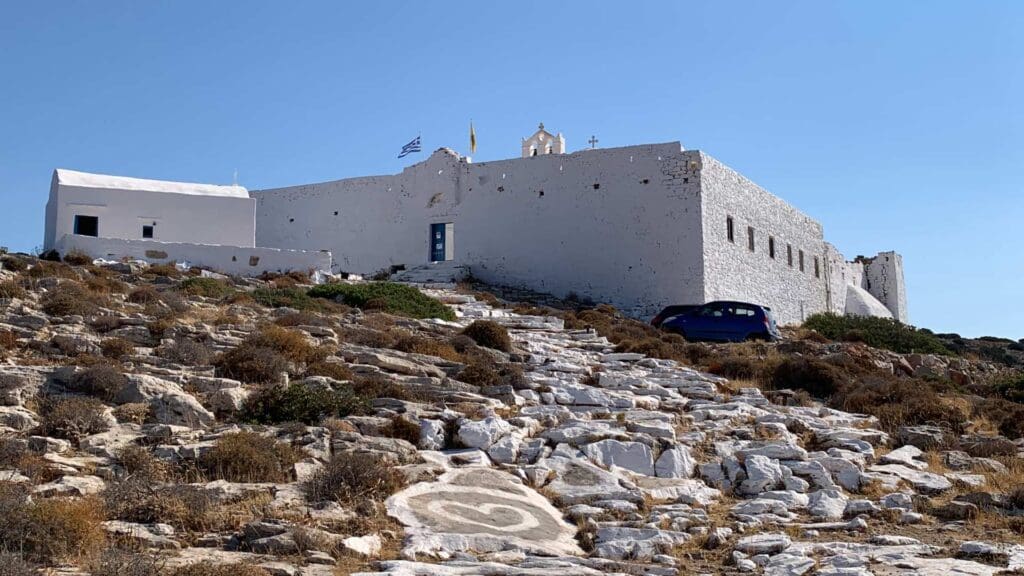 A white stone building perched on Sikinos Island features a sloped pathway and dry vegetation. The Greek flag flutters proudly above.