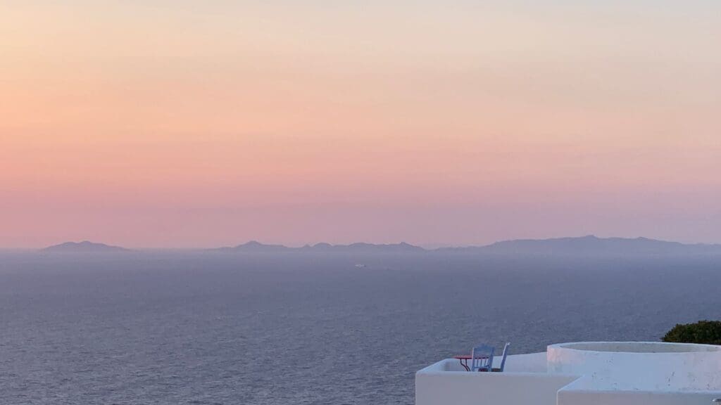 Sunset view over the sea with distant islands on the horizon, featuring a white building and a chair in the foreground, capturing the serene beauty of Sikinos Island.