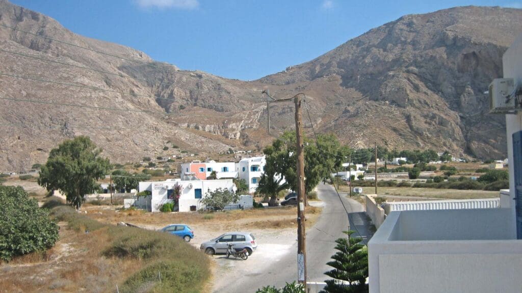 The serene rural landscape of Sikinos Island unfolds with a road lined by quaint parked cars and charming residential buildings, set against the backdrop of rugged mountains under a clear blue sky.