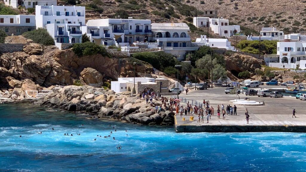 A coastal scene on Sikinos Island features people gathered on a dock near turquoise waters, surrounded by rocky cliffs and white buildings in the background.
