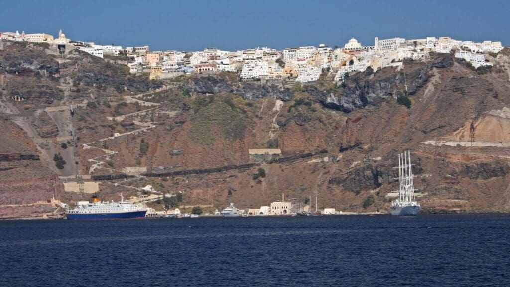 A hillside town with white buildings perches above a rocky cliff, reminiscent of Santorini. Nearby, two ships are docked at the ferry port by the shoreline, embraced by clear blue water.