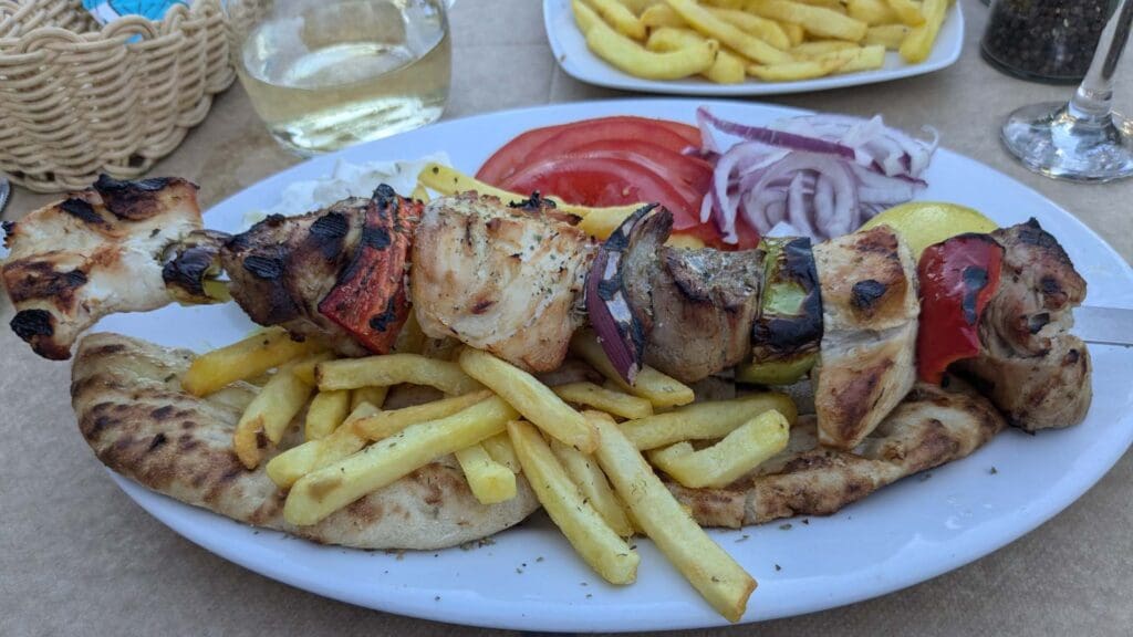 Savor the flavors of Greek street food with grilled meat and vegetables on skewers, served alongside pita bread, crispy fries, juicy tomato slices, and golden onion rings on a white plate.