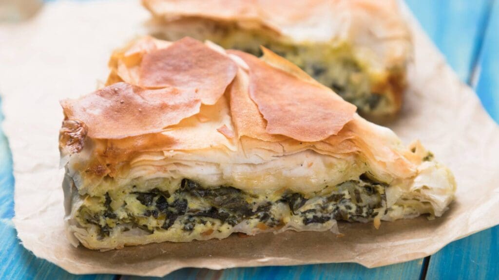 A slice of spinach and cheese pie with flaky pastry layers, reminiscent of Greek street food, rests invitingly on parchment paper.
