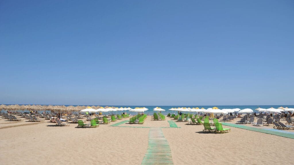A sandy beach with rows of green sun loungers and straw umbrellas under a clear blue sky captures the essence of Heraklion beaches.