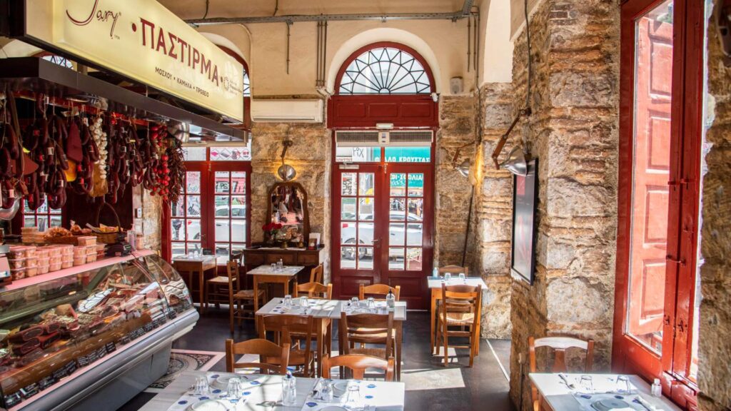 Step into the interior of this rustic Athens restaurant, adorned with stone walls and wooden tables. Large windows let in natural light, creating a cozy atmosphere perfect for Christmas in Athens. On the left, a display case showcases delightful deli products.