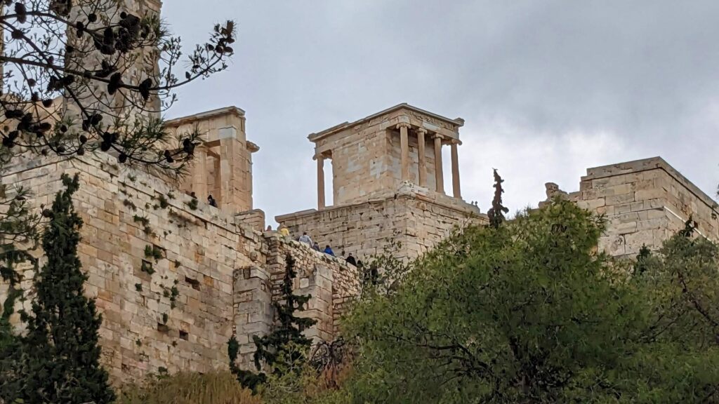 The ancient stone building with columns, reminiscent of the Acropolis, stands majestically on a cloudy day, surrounded by trees.