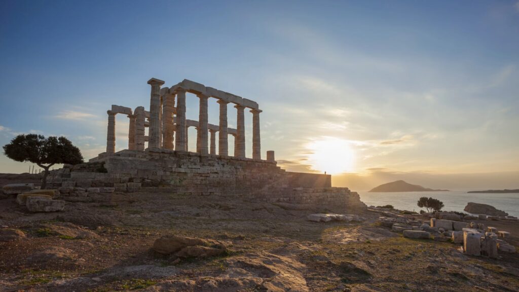 Ancient temple ruins with columns stand majestically at sunset, their silhouette etched against the rocky coast and calm sea. As the distant hills embrace the horizon, one might wonder how many days in Athens it takes to truly absorb such timeless beauty.